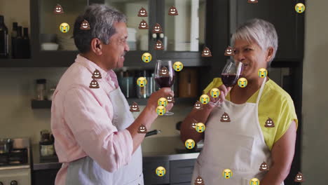 animation of emoji icons over senior biracial couple drinking wine in kitchen