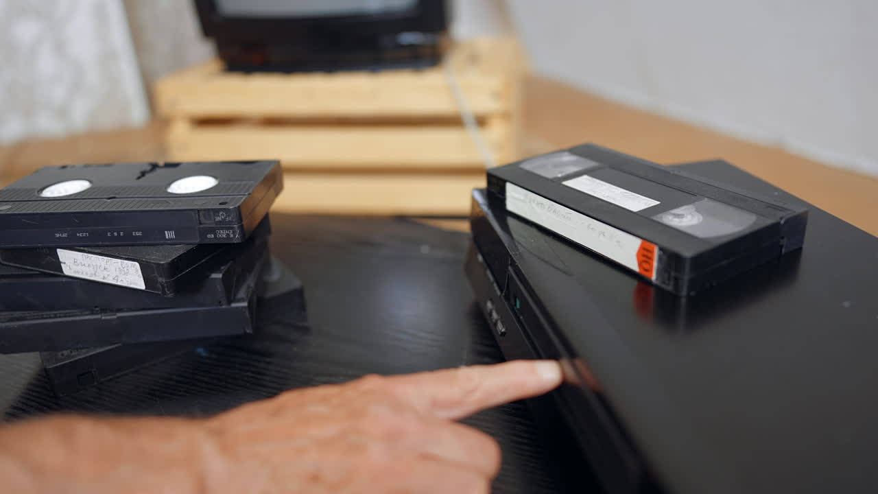 A hand takes a VHS tape from the pile and shoves into the player. Close up. Screen at blurred backdrop