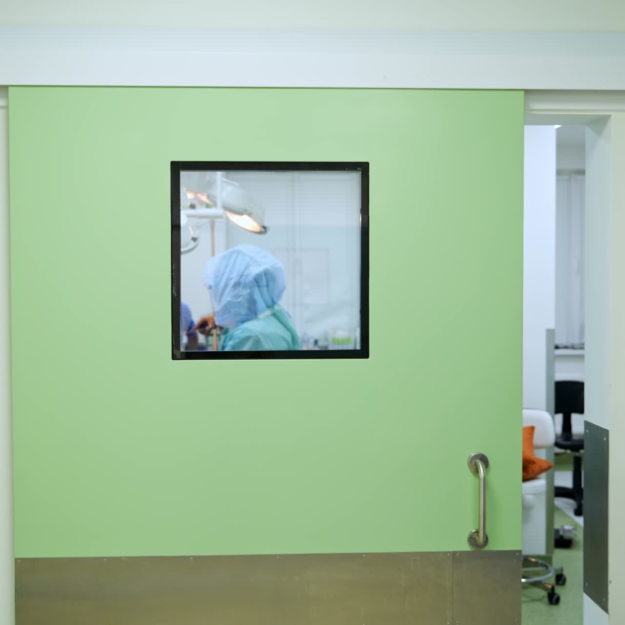 Square window in a door to a light modern surgery room. Doctor in protective suit applies iodine on the patient. Female nurse leaves the room