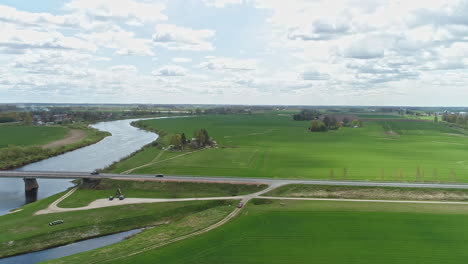 Aerial-drone-backward-moving-shot-over-green-farmlands-and-gardens-beside-a-river-with-cars-passing-by-over-a-bridge-during-evening-time