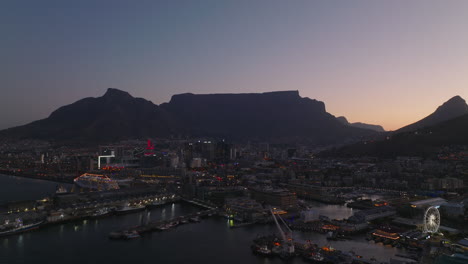 Fly-above-city-marina-at-sea-coast.-Buildings-and-streets-of-evening-town-and-silhouette-of-mountains-against-sky-at-dusk.-Cape-Town,-South-Africa