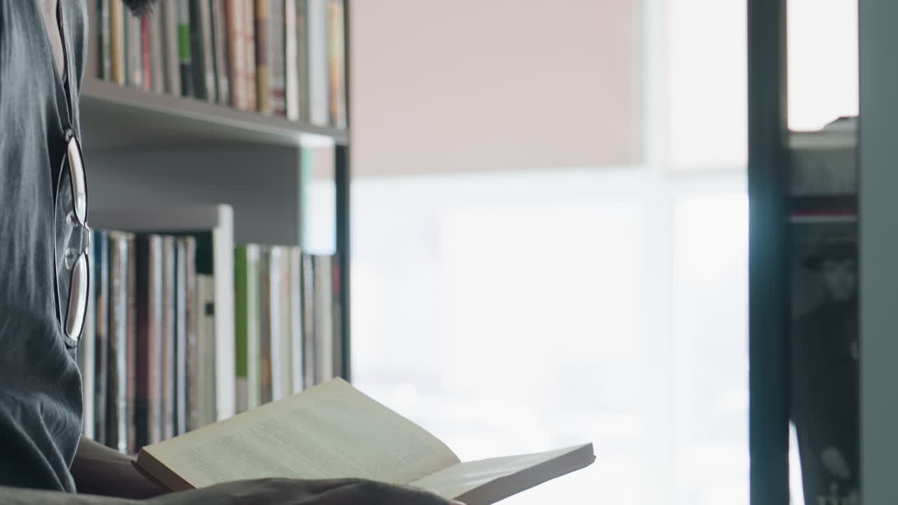 Giovane uomo visto da dietro in piedi in una biblioteca, che studia vicino agli scaffali con la luce soffusa del giorno che filtra attraverso la finestra, indossa un berretto e una camicia casual