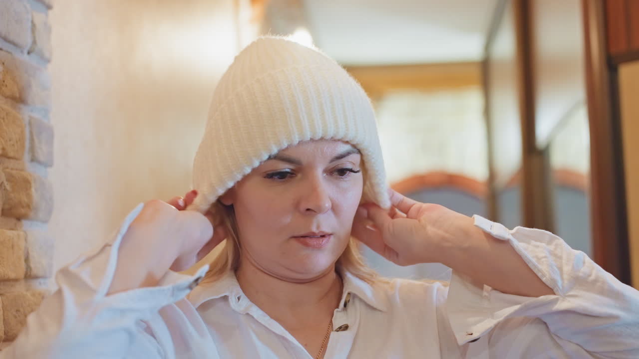 Caucasian woman trying beanie in hallway mirror, adjusting knit hat while checking reflection. Soft warm lighting, brick wall texture, wooden doorway, casual white shirt, relaxed morning routine