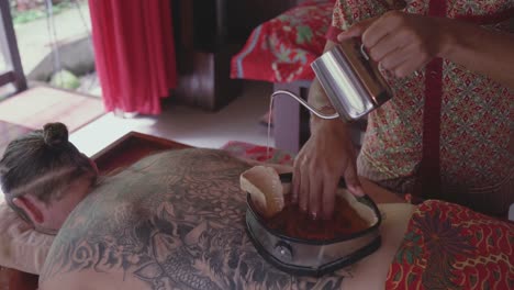 spa therapist pours warm oil into a wax-lined container on the back of a male patient enjoying a kati vasti ayurvedic treatment and stirs it with his hand