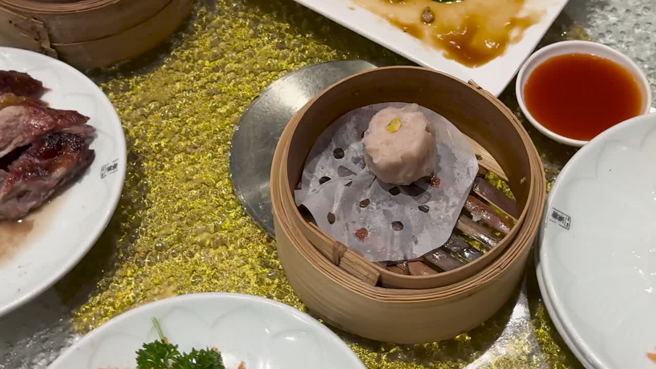 A single steamed dumpling in a bamboo steamer rotates past bok choy and sauces