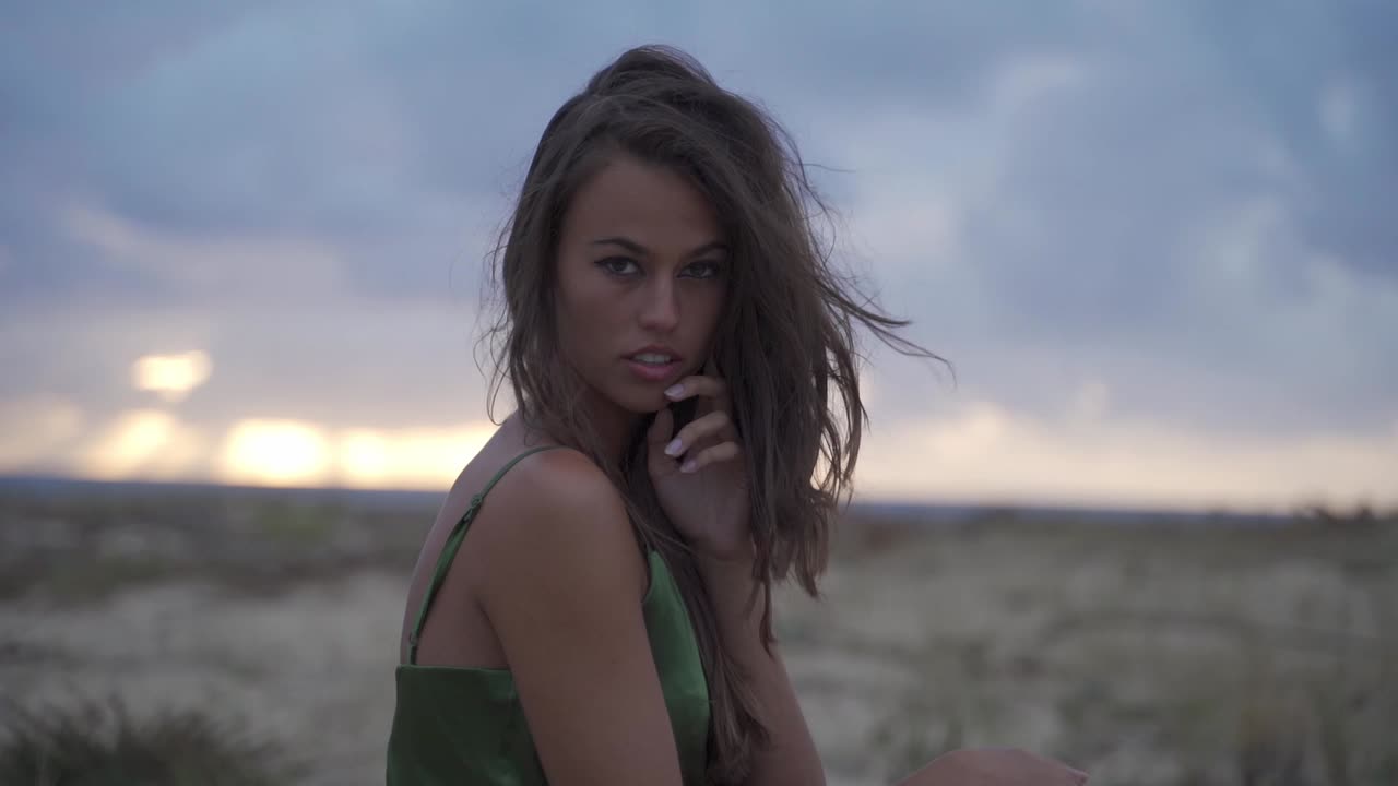 mujer de cabello oscuro con una mirada seductora, poderosa y segura de sí misma, con un vestido verde elegante en la playa al atardecer nublado y el viento soplando a través de su cabello