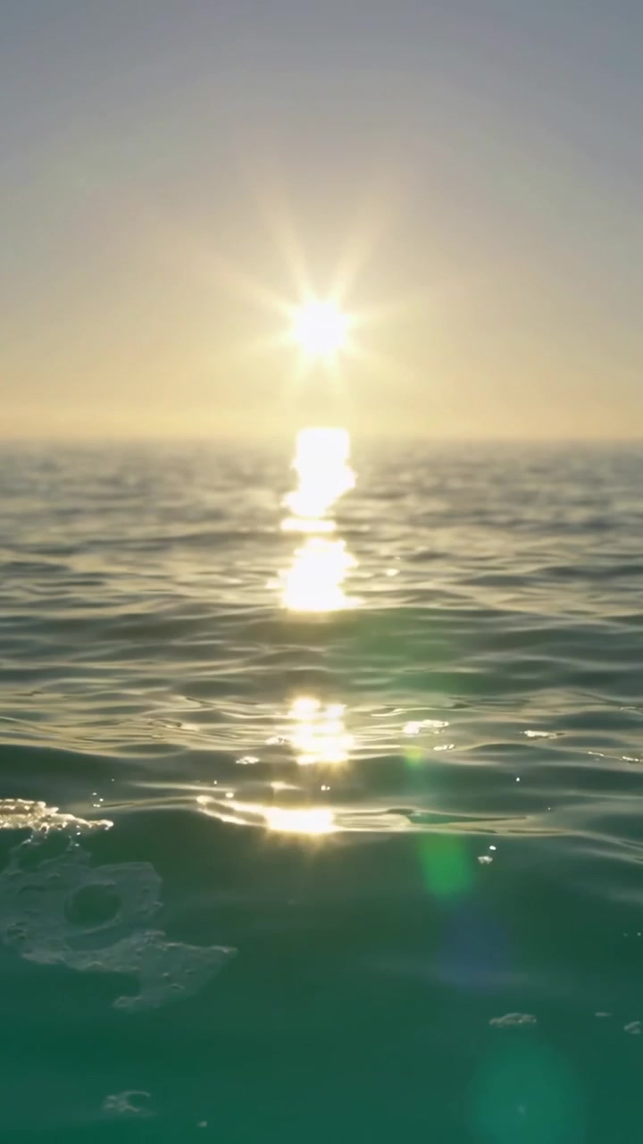 Vertical video: Near horizon sun casting long reflection on ocean, foam patches, green flare moving