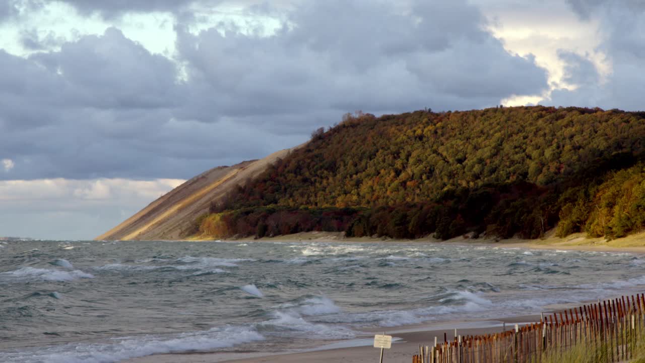 Potężne fale jeziora Michigan rozbijają się pod pochmurnym jesiennym niebem, a w tle wznoszą się wydmy Sleeping Bear Dunes, tworząc dramatyczny, szeroki i stabilny kadr jesiennej linii brzegowej.