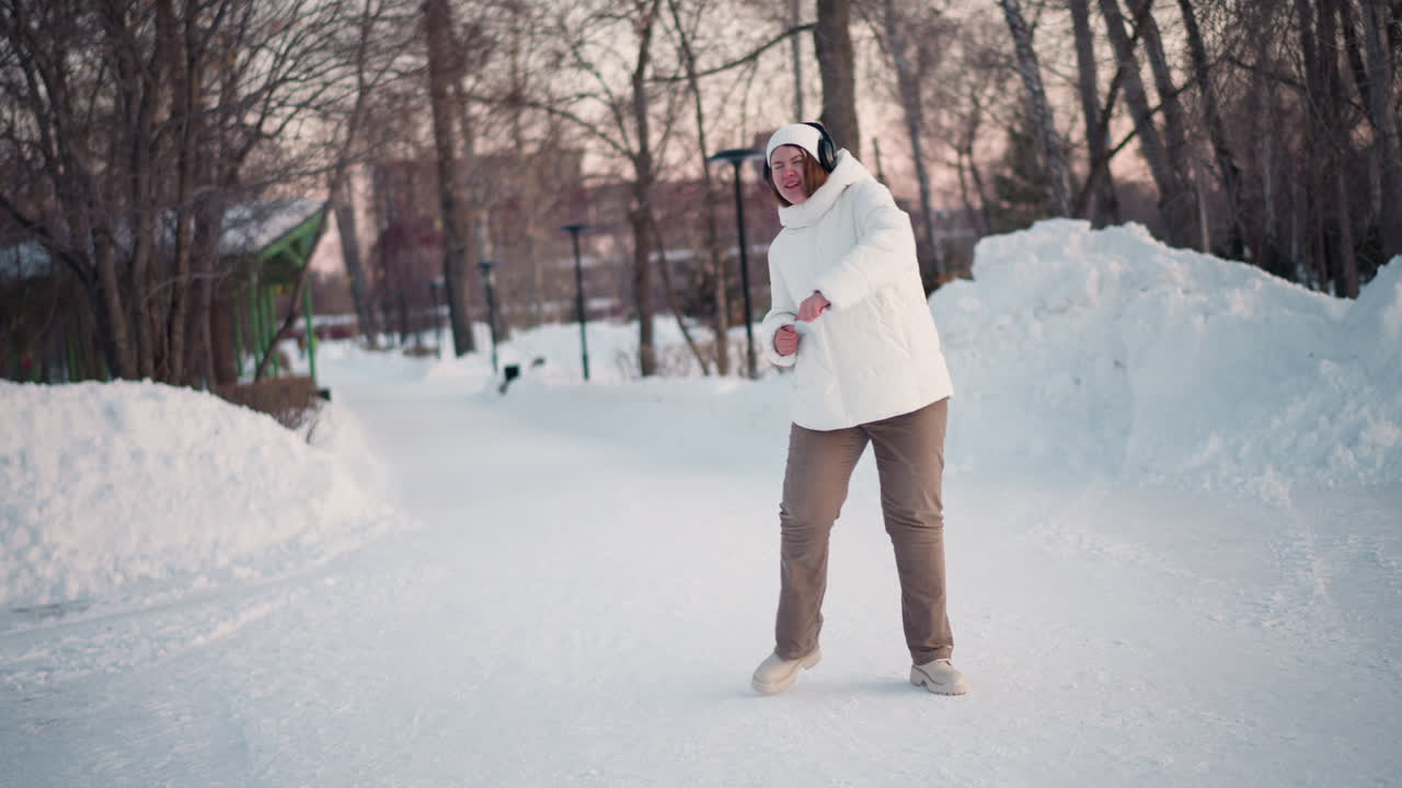 Creative wearing white beanie and headset dances joyfully on snowy path at sunset in puffer coat and tinted goggles, swaying body to music with bare winter trees and warm glow in background