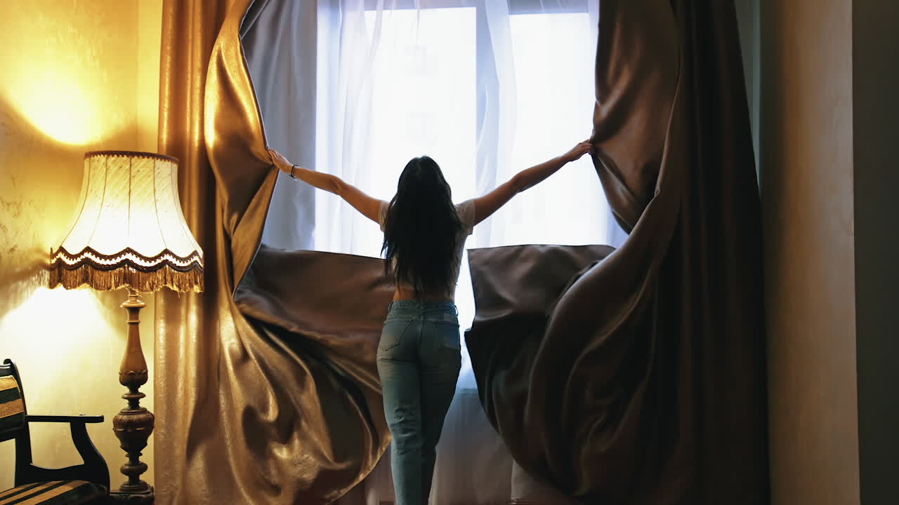 Happy optimistic young woman opens window. Smiling girl running happily to the window and opening dark curtains in luxury hotel room.