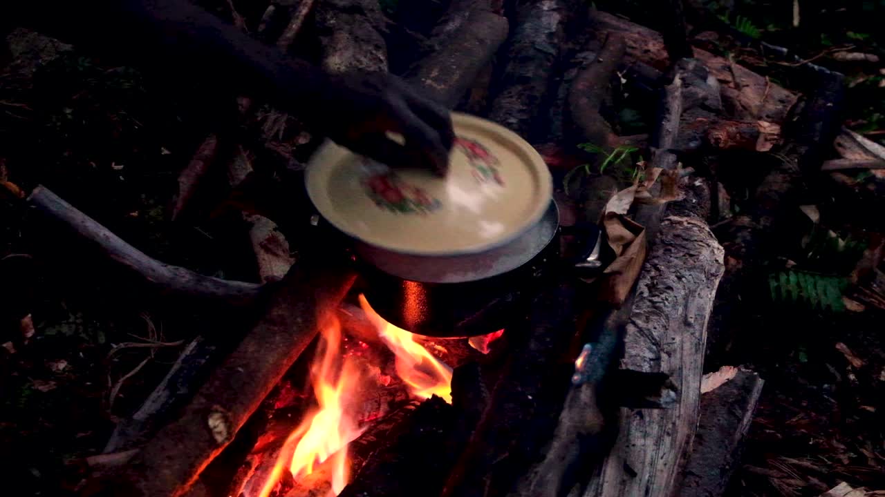 Cooking dinner on overnight walk hike in the remote wilderness and rainforest. Hand putting lid on hot saucepan pot cooking on open log fire with flames and logs