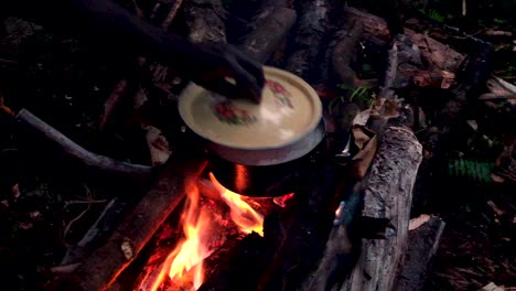 cooking dinner on overnight walk hike in the remote wilderness and rainforest. hand putting lid on hot saucepan pot cooking on open log fire with flames and logs