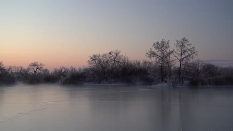 Dampf,-Der-Von-Einem-Zugefrorenen-See-Mit-Bäumen-Aufsteigt,-Die-Vom-Sonnenaufgang-Silhouettiert-Werden