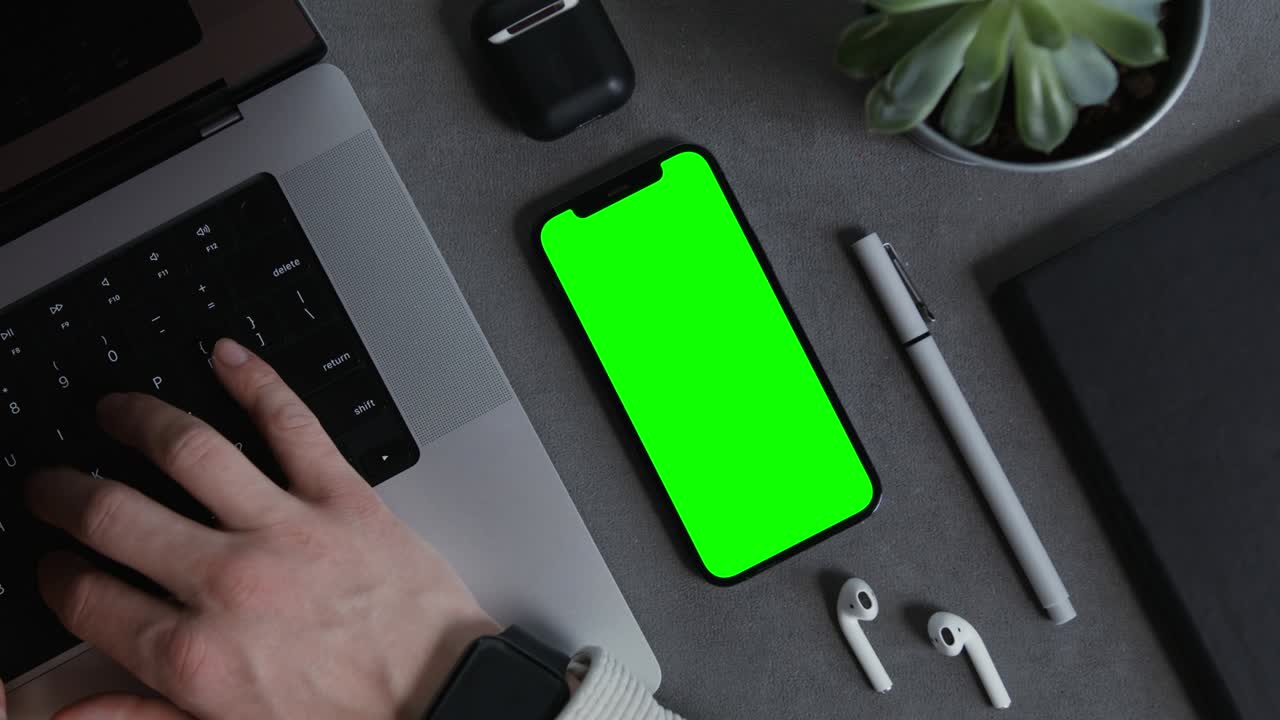 un teléfono móvil con una pantalla verde se encuentra en el escritorio del hombre escribiendo en una computadora portátil