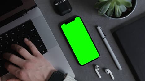 a mobile phone with a green screen lies on the man's desktop typing on a laptop