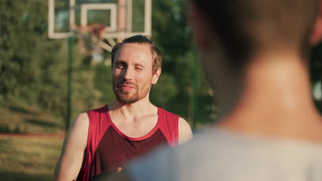 Retrato-De-Un-Apuesto-Jugador-De-Baloncesto-Sonriente-Sosteniendo-Una-Pelota-Y-Explicando-Algo-A-Su-Amigo,-Parado-Frente-A-él-En-Un-Primer-Plano-Borroso