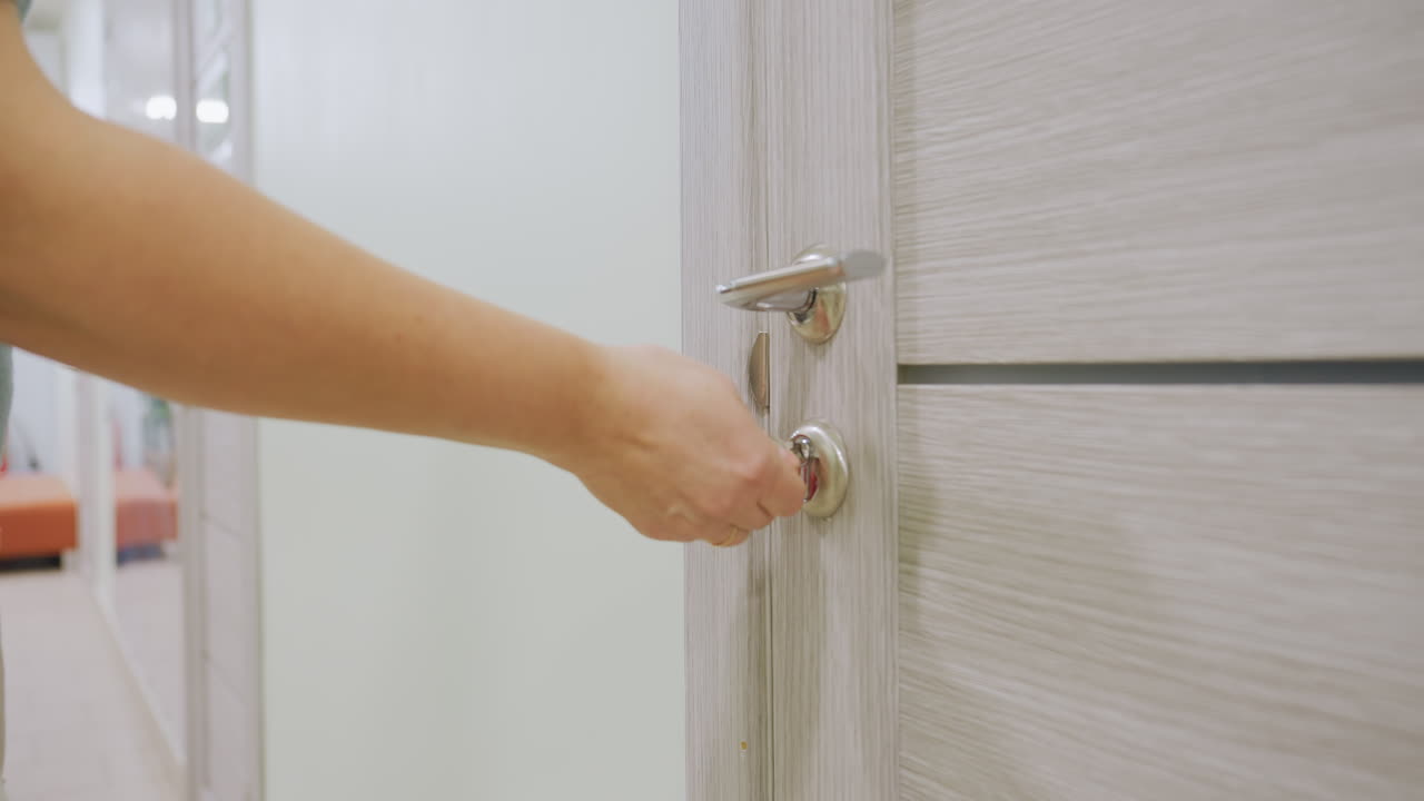 Close up of fair female hand unlocking wooden door with metallic handle and closing it behind her as hallway mirror reflects faint movements of people outside suggesting subtle indoor activity