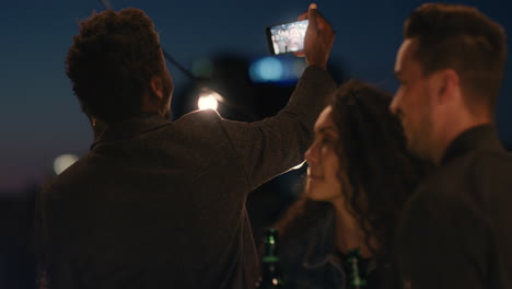 Amigos-Felices-De-La-Fiesta-En-La-Azotea-Divirtiéndose-Joven-Afroamericano-Tomando-Fotos-Usando-Un-Teléfono-Inteligente-Compartiendo-El-Estilo-De-Vida-Del-Fin-De-Semana-En-Las-Redes-Sociales-Por-La-Noche