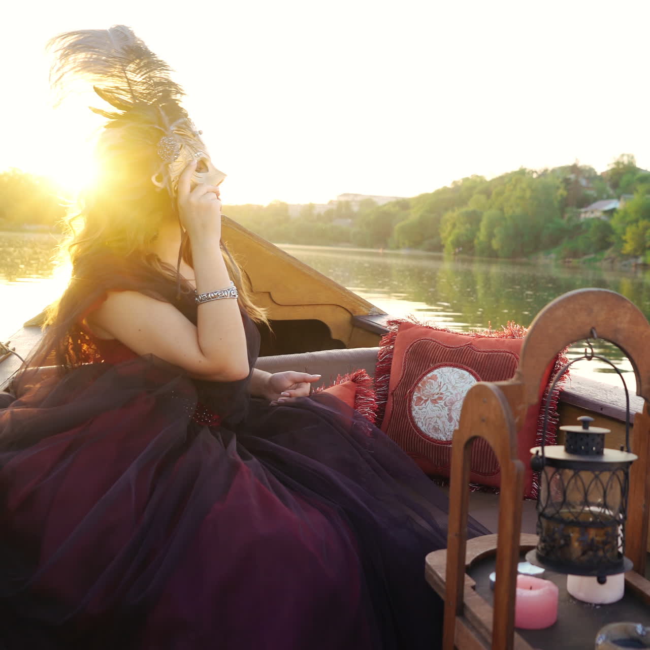 Gorgeous female in masquerade costume sitting in gondola and looking at beautiful natural landscape. Woman holding carnival mask floating in a boat at sunset.
