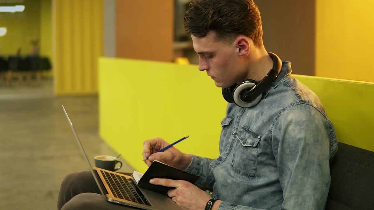 Attractive sexy male in his 20's sitting in a well lit indoors space on sofa, writing down something in his notepad with laptop on his laps.