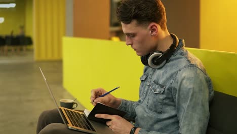 attractive sexy male in his 20's sitting in a well lit indoors space on sofa, writing down something in his notepad with laptop on his laps.