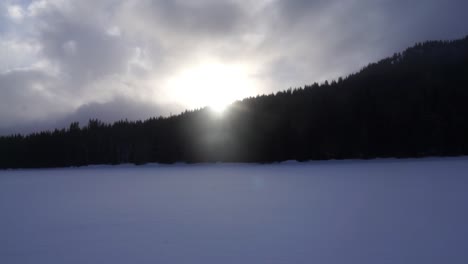 Snow-blowing-across-frozen-lake-at-sunset-with-trees-in-background