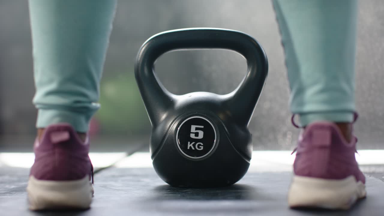 Mujer afroamericana adopta una postura en la colchoneta, sujetando una pesa rusa de 5 kg y entrenando.