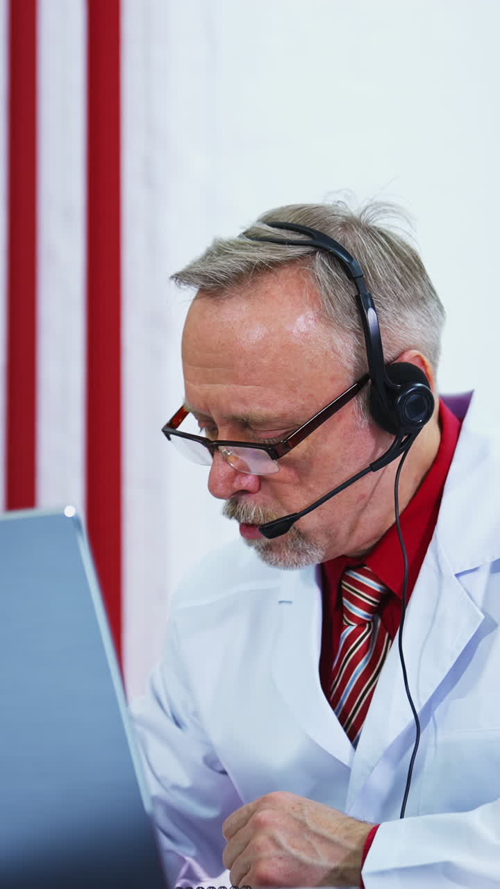 Telemedicine. Old physician listens to a patient from laptop. Male doctor wear headset make online video call consultation to a patient in clinic. Vertical video