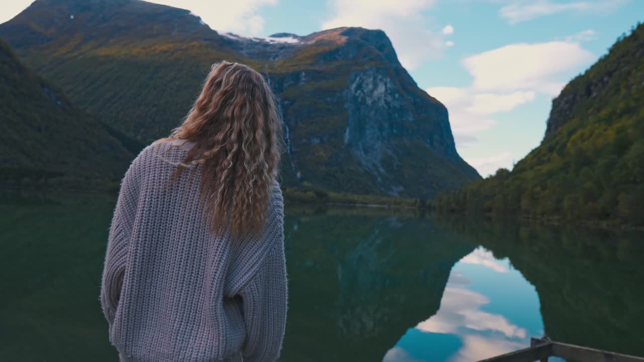 Blonde girl wearing a white sweater, observing beautiful scenic view of a large lake and mountains in the background