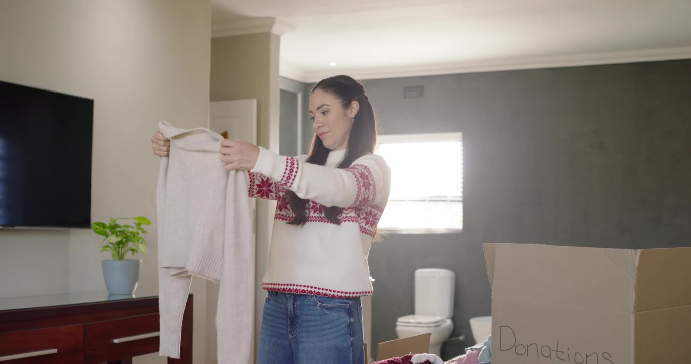 Adult woman picking up sweater from pile, inspecting and folding before placing into Donations box