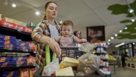 A-little-baby-girl-sits-in-a-cart-and-looks-around-everything-during-her-shopping-with-her-mother-in-the-supermarket