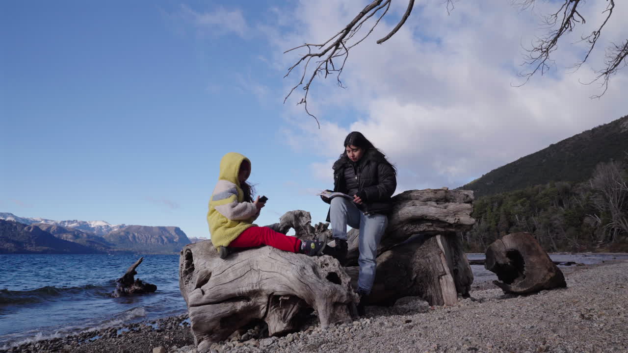 Two girls sit on large driftwood, one with a book, the other on her phone, surrounded by calm water