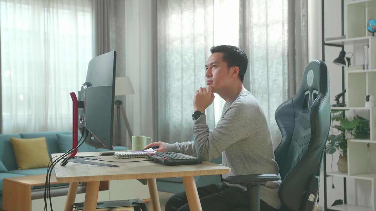 Asian Man In Long Sleeved T-Shirt And Black Pants Thinking Then Typing On Desktop Computer While Working At Home.
