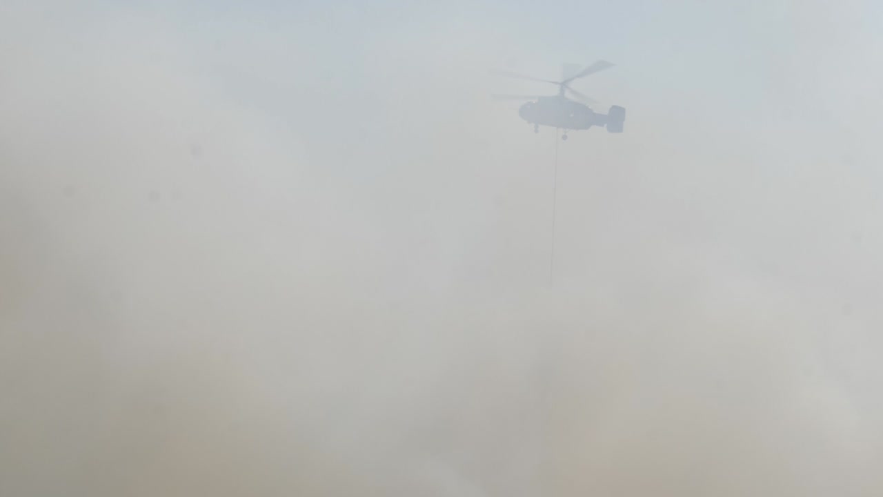 un helicóptero de extinción de incendios emerge del espeso humo de los incendios forestales con un cubo de agua para ayudar a extinguir las llamas.