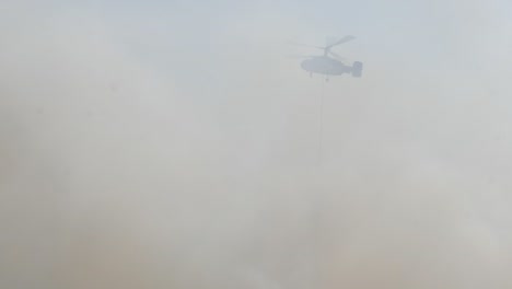 a fire fighting helicopter emerges from thick forest fire smoke with a water bucket to help extinguish the flames