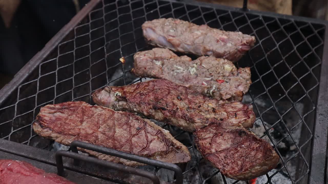 Raw and seared steaks are being grilled on a metal grate over hot charcoal, with a gloved hand using tongs to turn the meat