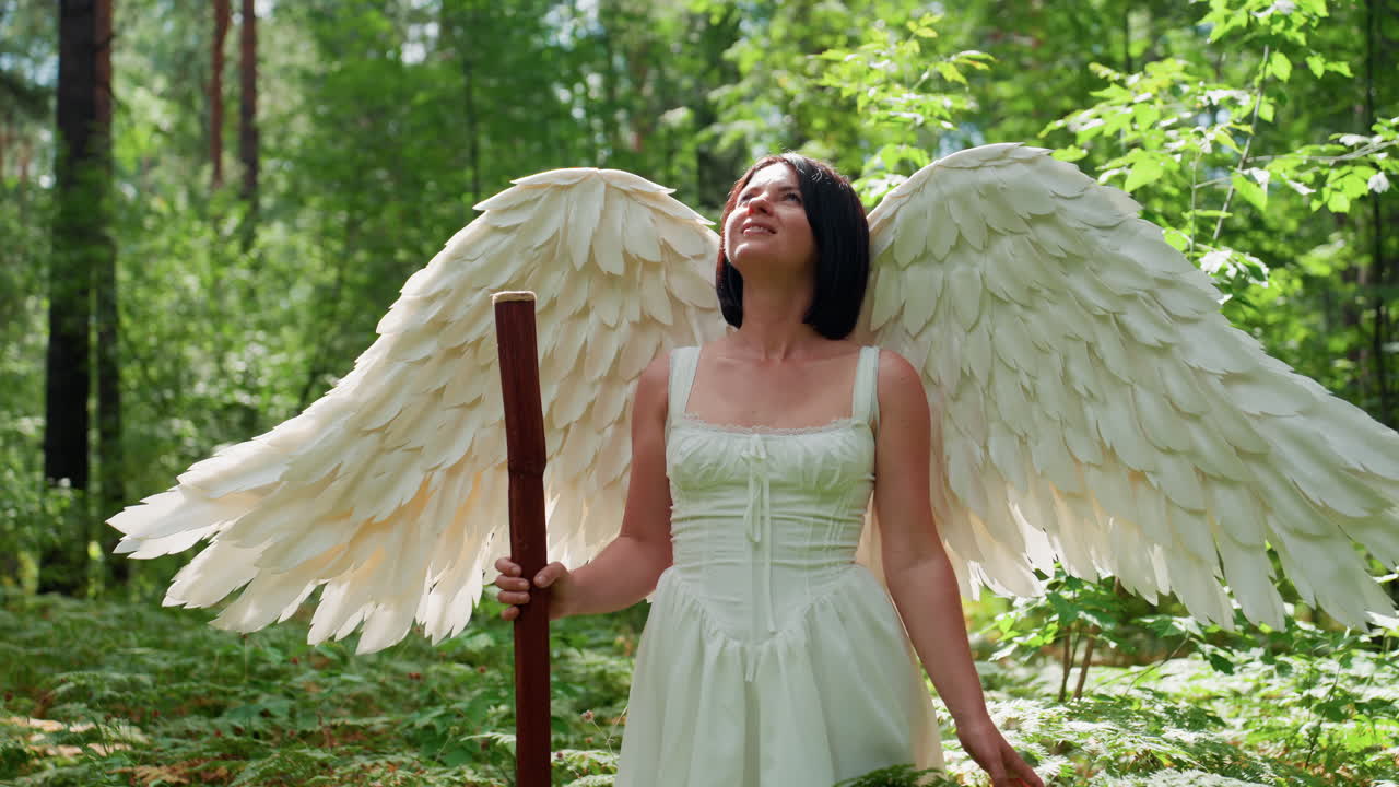 Magical creature with large white wings and wooden staff standing in lush green forest bathed in sunlight observing surroundings with calm expression symbolizing purity