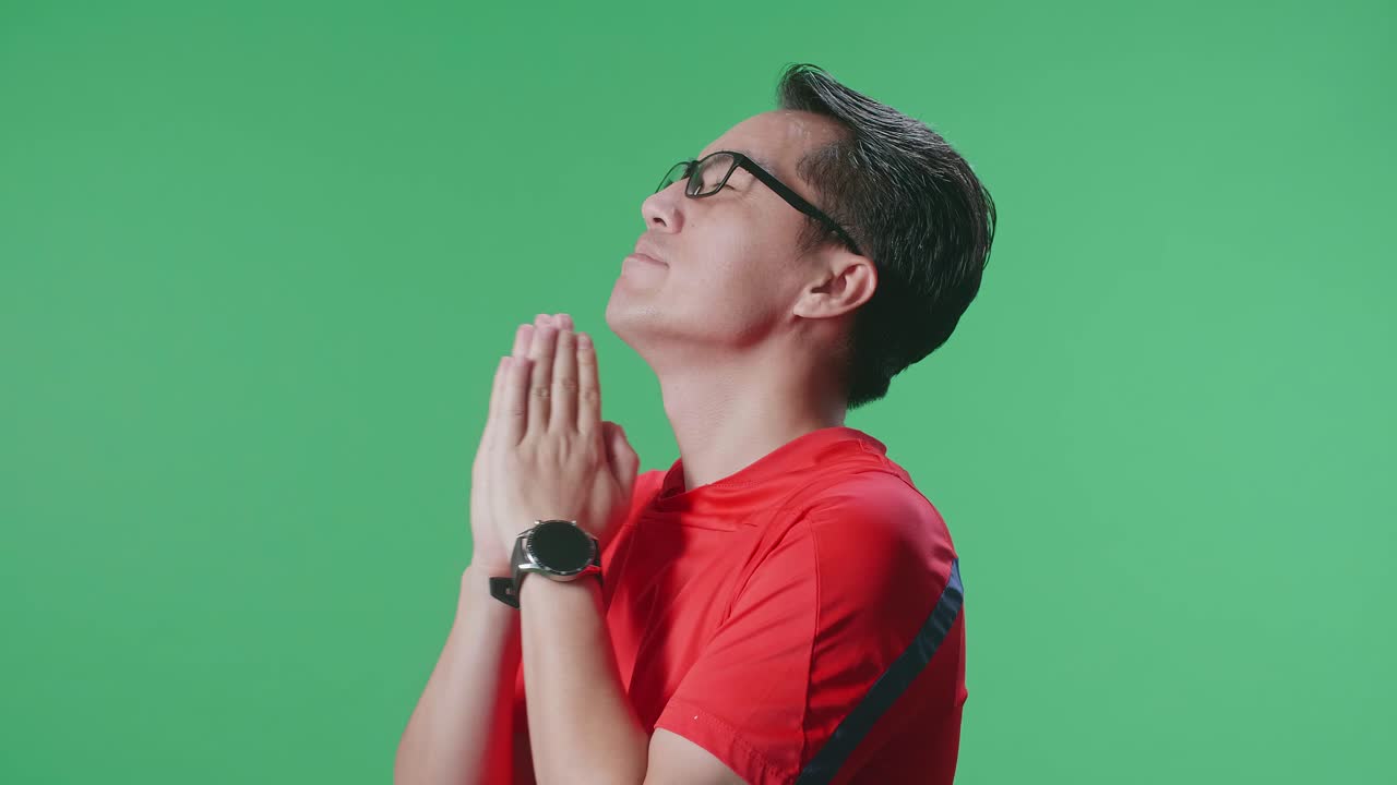 눈을 닫은 으로 쳐다보는 모습: 공을 들고 있는 아시아인 남자가 팀을 위해 기도하다가 손을 들고 초록색 스크린 배경에서 축구 경기를 응원하고 있다.