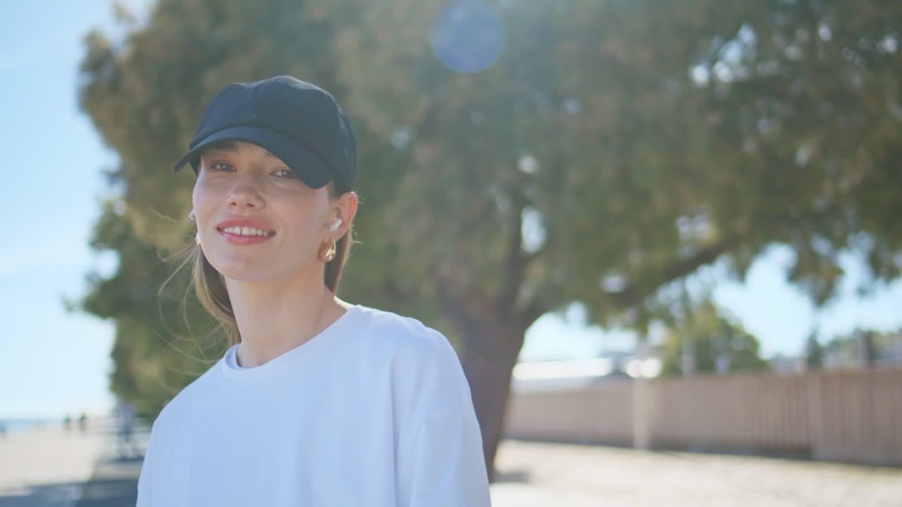 Carefree woman adjusting earphones standing riverside in black cap closeup