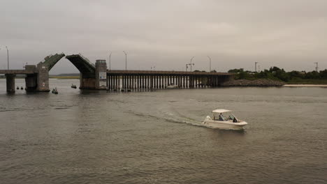 a low angle aerial view of a small boat sailing on baldwin bay, past a draw bridge opening on a cloudy day