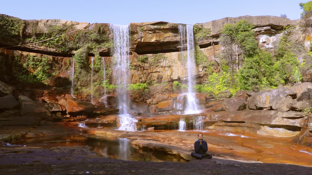 joven meditando en la cascada natural que cae desde la cima de la montaña en el día desde un video de ángulo bajo tomado en phe phe fall meghalaya india