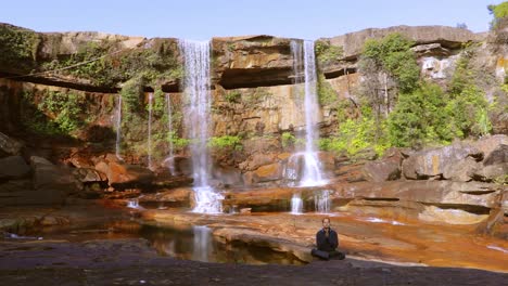 Junger-Mann-Meditiert-über-Den-Natürlichen-Wasserfall,-Der-Tagsüber-Von-Der-Bergspitze-Fällt,-Aus-Einem-Video-Aus-Niedrigem-Winkel,-Das-Bei-Phe-Phe-Fall-Meghalaya-Indien-Aufgenommen-Wurde