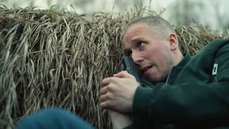 a close view of a man in a green jacket lying against dry grass, holding a pistol, and looking tense