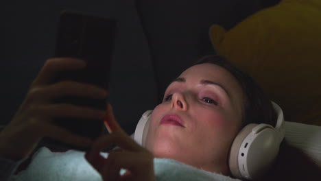 mujer con auriculares inalámbricos acostada en el sofá en casa por la noche transmitiendo música o viendo una película en el teléfono móvil 3