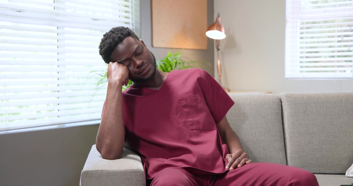 African American man in maroon scrubs scrolling phone on gray sofa during break, drifting to sleep