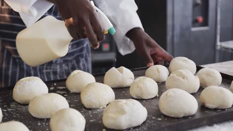 african american male baker working in bakery kitchen, spraying rolls in slow motion