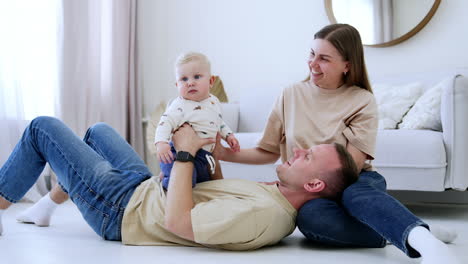 smiling caucasian man lies on the floor his head on wife's knees. dad holds a plump baby boy sitting on his belly. happy family at home.