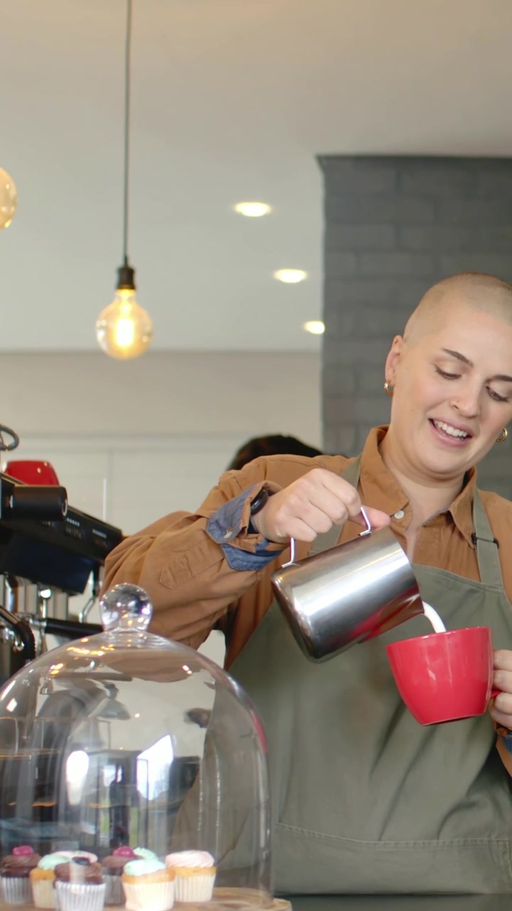Video verticale: Barista con grembiule verde che versa il latte in una tazza rossa mentre un cliente fa una domanda al bar.