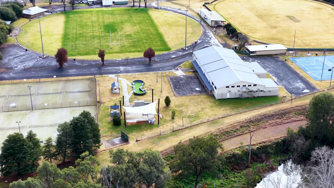Drone footage glides over a small town sports complex with green fields, tennis courts, playground, and community buildings under bright daylight