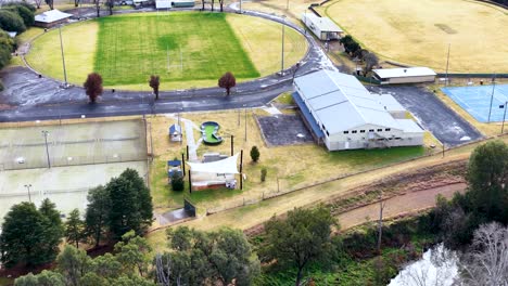 drone footage glides over a small town sports complex with green fields, tennis courts, playground, and community buildings under bright daylight